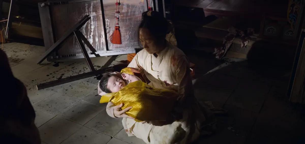 Escena de una mujer sosteniendo a un bebé envuelto en tela amarilla, como se ve en 'Kingdom' de Netflix.