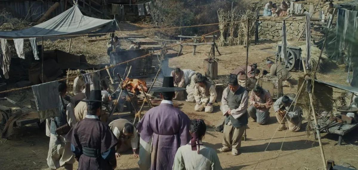 Una reunión de personas, algunas arrodilladas, alrededor de un asado al aire libre, como se muestra en 'Kingdom' de Netflix.