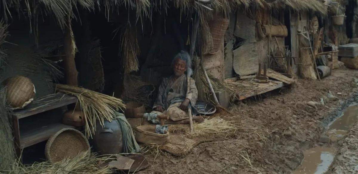 Una aldea con una persona sentada frente a una cabaña rudimentaria, escena de 'Kingdom' de Netflix.