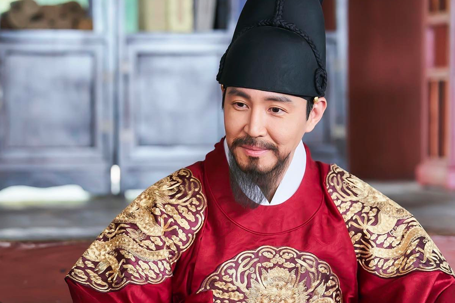 劇中王身著紅色王室服裝坐在宮殿中，傳達韓國古裝劇中的王的形象，來自於《王后傘下》劇照。