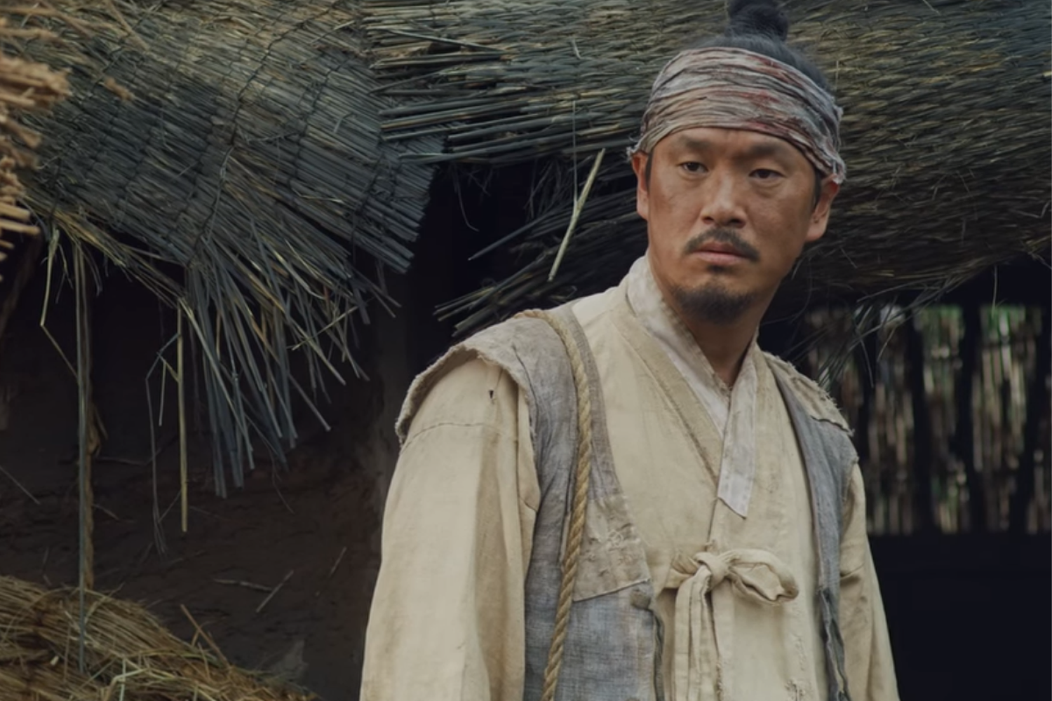 《屍戰朝鮮》中常民穿著淺色衣服坐在麥草堆旁，背景是簡陋的稻草屋。