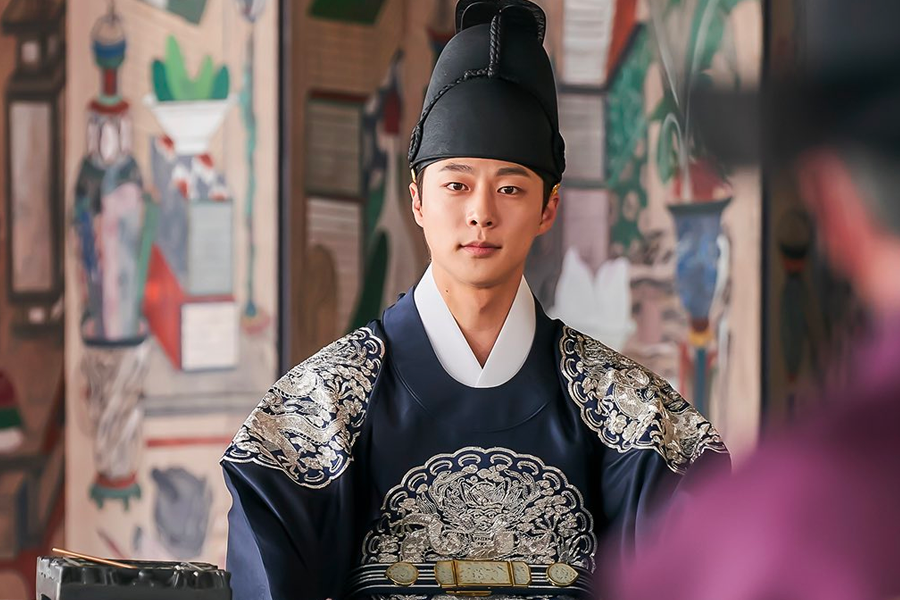 韓國古裝劇《王后傘下》中，世子身穿藍色宮服，坐在宮殿內的場景，具有朝鮮宮廷的氛圍。