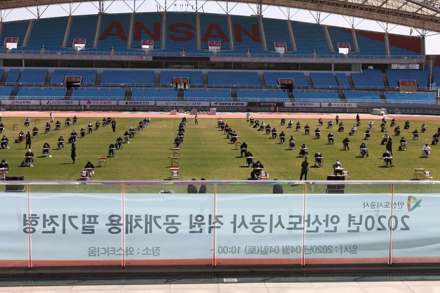 韩国，一个室外足球场上进行的笔试场景，考生们在草地上相隔一定距离，统一佩戴口罩以保持社交距离。
