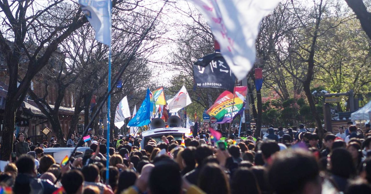 一场文化活动中，道路被旗帜及参与者的集会所填满，部分旗帜展示了彩虹图案。