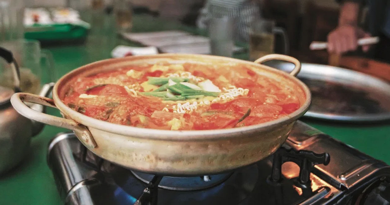 Hình ảnh món canh Budae Jjigae Hàn Quốc đang nấu trên bếp, nổi bật với sắc cam đỏ của nước dùng cùng mì, xúc xích, và các loại rau. Bầu không khí ở một quán ăn thường ngày tại Hàn Quốc.