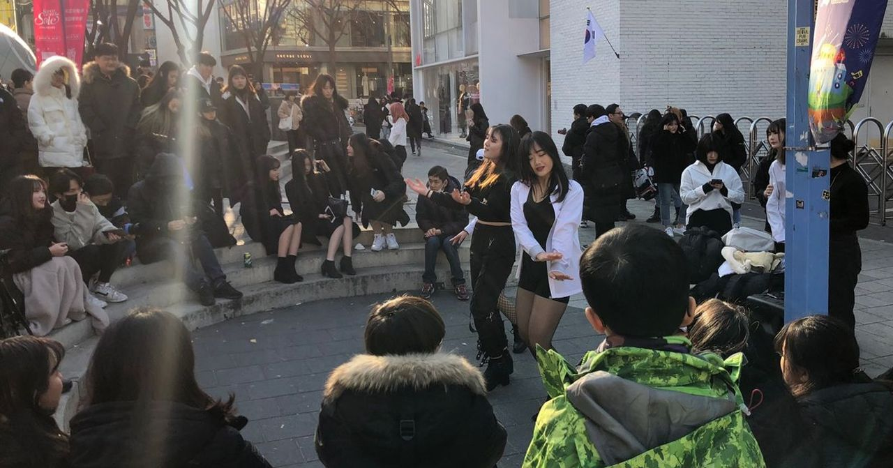 首爾商圈街頭上，年輕人圍觀街頭表演者的場景，顯示韓國流行文化和社會生活的一部分，吸引路人的舞蹈表演。