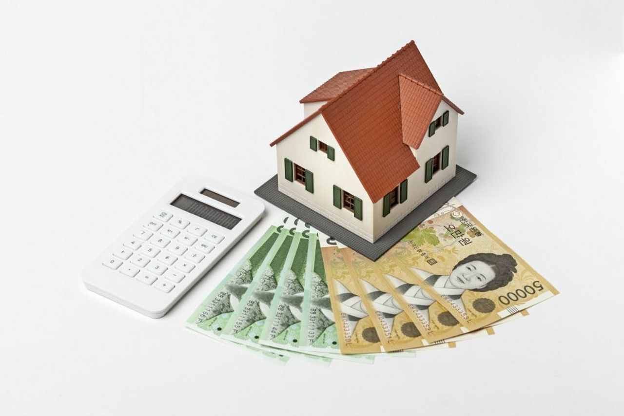 桌上擺放著模型小屋、現金和計算機，象徵著房地產市場、金融和經濟的各種考量與計算。