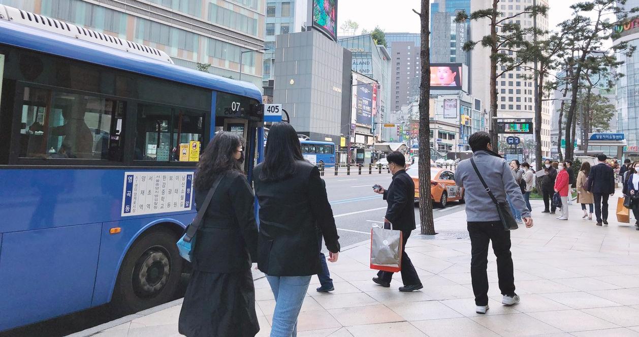 Khung cảnh bên ngoài tại bến xe buýt gần Lotte Department Store ở Seoul, Hàn Quốc. Nhiều người đi bộ và xe buýt số 405 sẵn sàng cho hành trình tiếp theo.