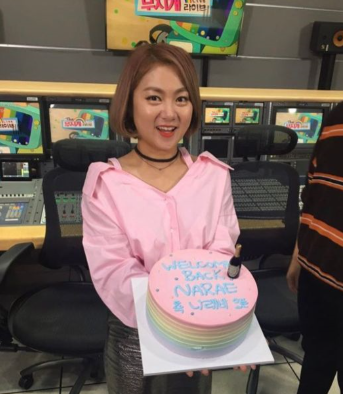 カラフルなホールケーキを持ってスタジオの前に立つ人。ケーキには「WELCOME BACK, NARAE」と書かれている。