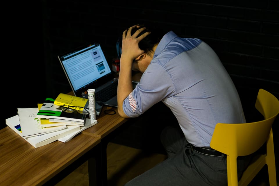 ชายคนหนึ่งนั่งบนเก้าอี้สีเหลืองข้างโต๊ะที่มีหนังสือกองอยู่ ด้านหน้ามีโน้ตบุ๊กที่เปิดเว็บไซต์เกี่ยวกับผลกระทบจากโควิด-19 เขาจับศีรษะด้วยมือทั้งสองข้างแสดงความเครียดและเศร้าใจในสภาพแวดล้อมการทำงานภายใต้สถานการณ์โรคระบาด