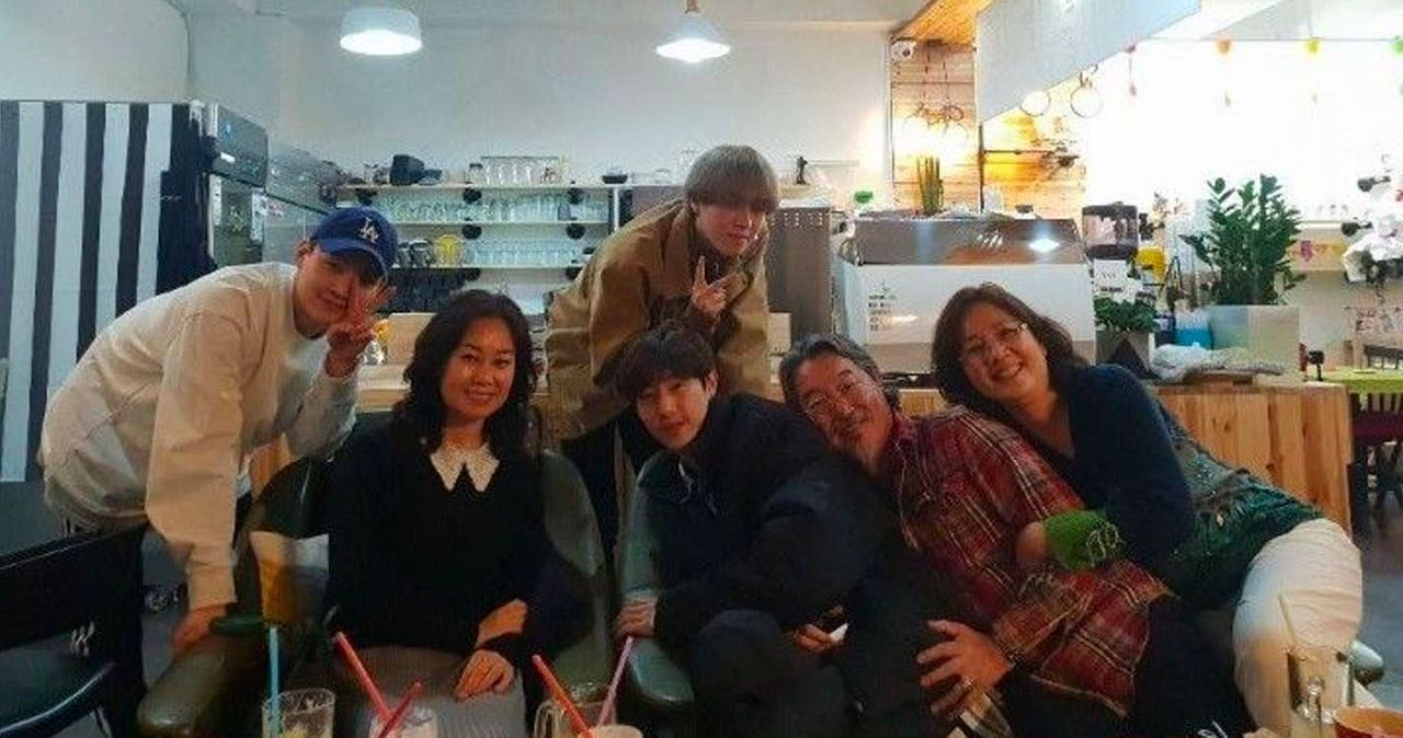 กลุ่มเพื่อนสนิทนั่งอยู่บนโซฟาในคาเฟ่ที่มีการตกแต่งด้วยแสงไฟที่อบอุ่น เบื้องหลังมีแถบเส้นสีขาวดำและชั้นวางของที่มีแก้วและกล่องหลายใบ