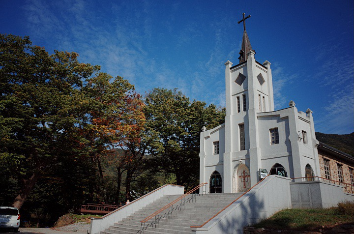 미리내 성지에서 촬영된 '사랑의 불시착' 장면으로, 예쁜 교회 건물이 있는 풍경입니다. 푸른 하늘과 주변의 나무들이 한층 더 아름다움을 더합니다. 이 장소는 따뜻하고 평화로운 분위기를 자아내며 극중 중요한 장면의 배경으로 사용되었습니다.