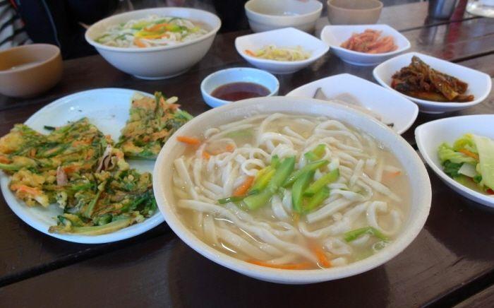 บะหมี่ร้อนกับเครื่องเคียงแบบเกาหลีในร้านอาหารพื้นบ้าน, สัมผัสรสชาติท้องถิ่นอย่างแท้ทรู (Hot noodles with traditional Korean side dishes served in a rustic setting).