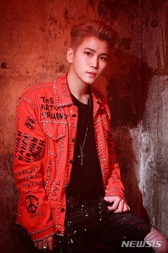 赤いジャケットを着たダンディ、韓国の有名作曲家でありプロデューサー、背景には壁が見える。ジャケットにはメッセージが書かれ、クロスネックレスをつけている。