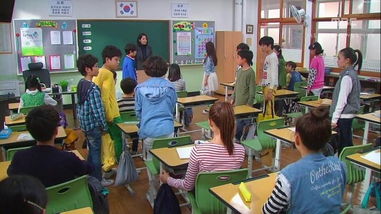 韓國學校的課堂場景，教室內學生站在課桌前，黑板上掛有韓國國旗。