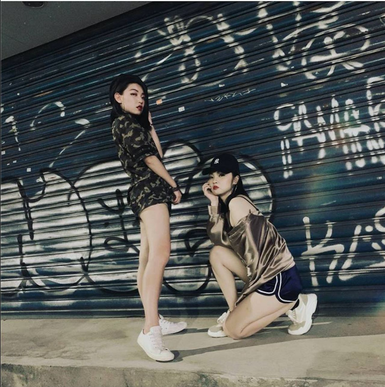 兩位陷阱妹在塗鴉鐵門前展示時尚穿搭，有oversize上衣和白色運動鞋等元素。