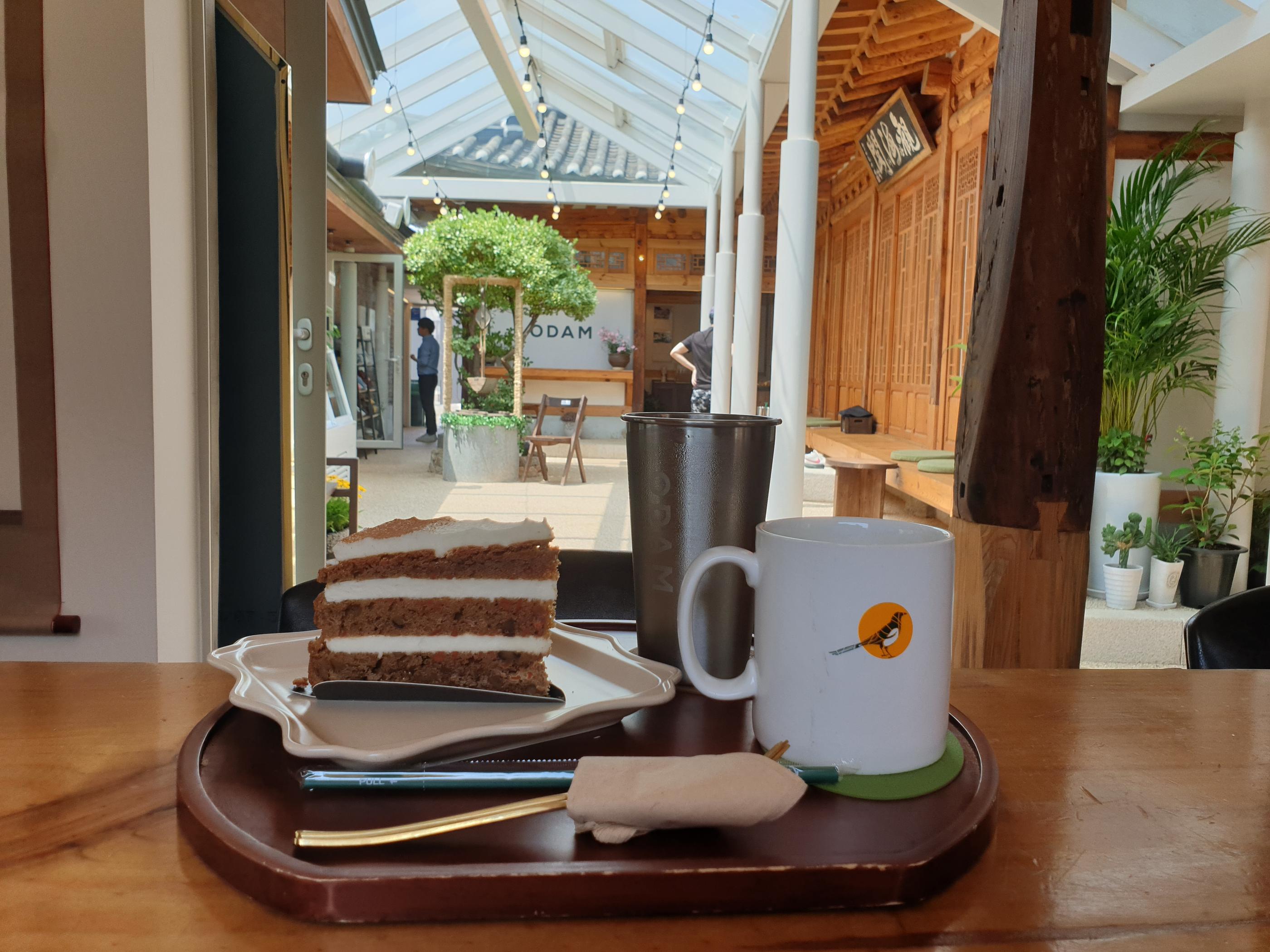 韓屋カフェで見えるキャロットケーキとホットドリンクのプレート、明るいテラスの中に心地よさを感じる場所。