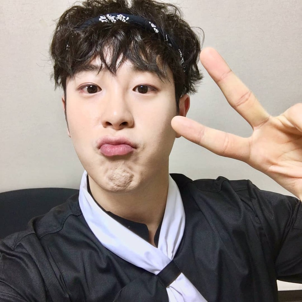 Block B成員P.O穿著黑色制服和白色領巾, 比出V字手勢, 表現輕鬆愉快