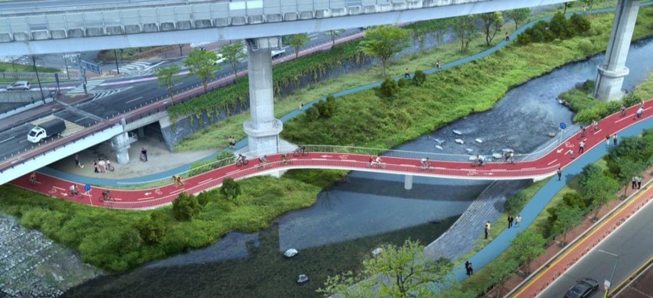 首尔市正在进行中的自行车道路建设项目，融合自然景观和现代桥梁设计。图中展示了一段红色自行车道沿着河流和桥梁延伸，体现出现代化城市交通和自然的结合。