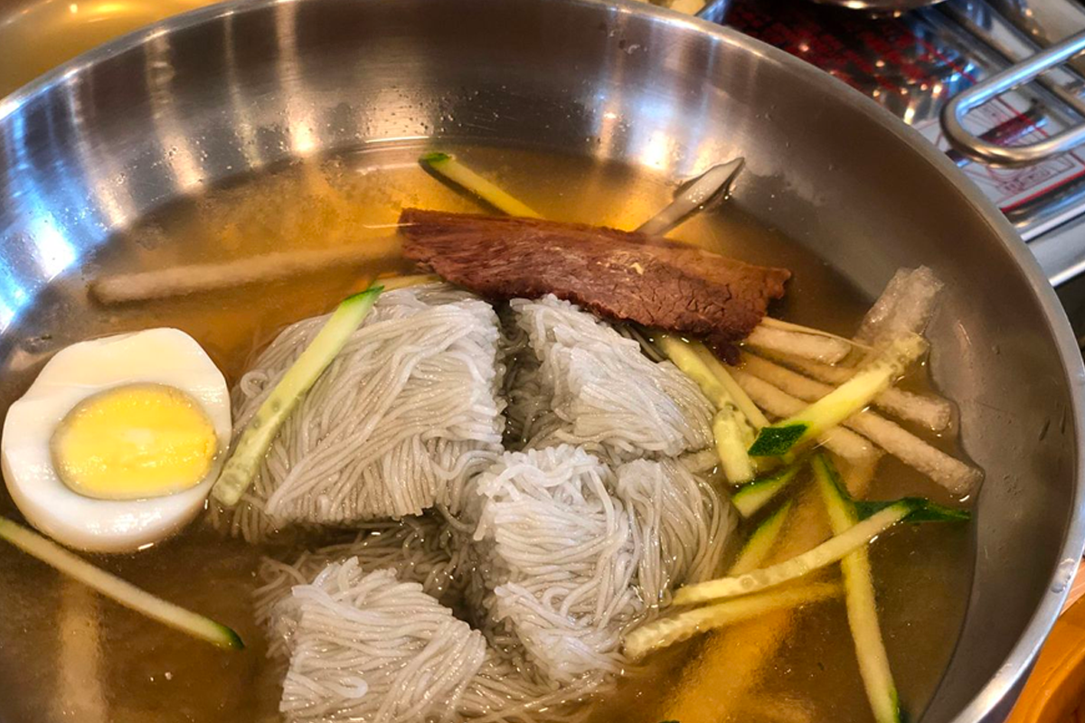 鋼盆中的韓國冷麵，湯汁清澈，加入半顆雞蛋、黃瓜條和牛肉片，展現夏日清爽美食形象。