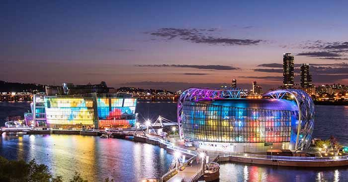 ทิวทัศน์สวยงามของสถานที่ท่องเที่ยวทางน้ำในโซลตอนเย็นพร้อมไฟสีสวยงาม บรรยากาศในภาพเป็นช่วงเวลาพระอาทิตย์ตกดิน บนแม่น้ำฮัน มีอาคารที่มีแสงไฟสวยงามซึ่งเป็นจุดท่องเที่ยวที่มีชื่อเสียง