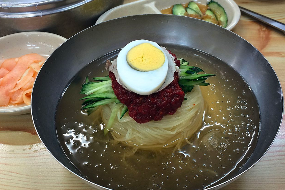 韓國小麥冷麵放在鋼盆裡，上面擺著黃瓜條、煮熟的雞蛋和細碎的牛肉，外觀清爽適合炎夏。
