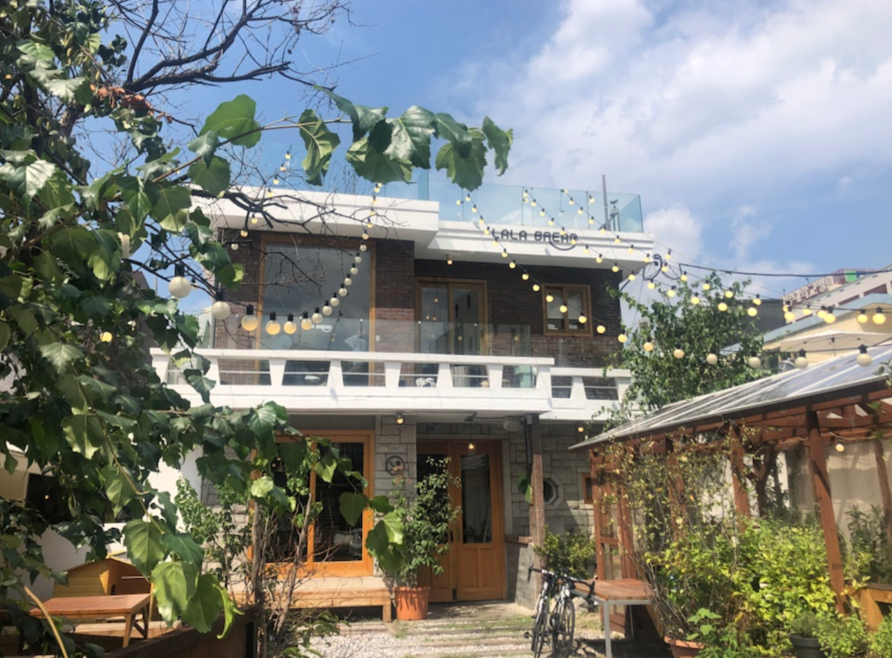 LALA BREADハンナム店の外観。緑の樹木と少し見える建物の一部、明るい空が背景にあります。