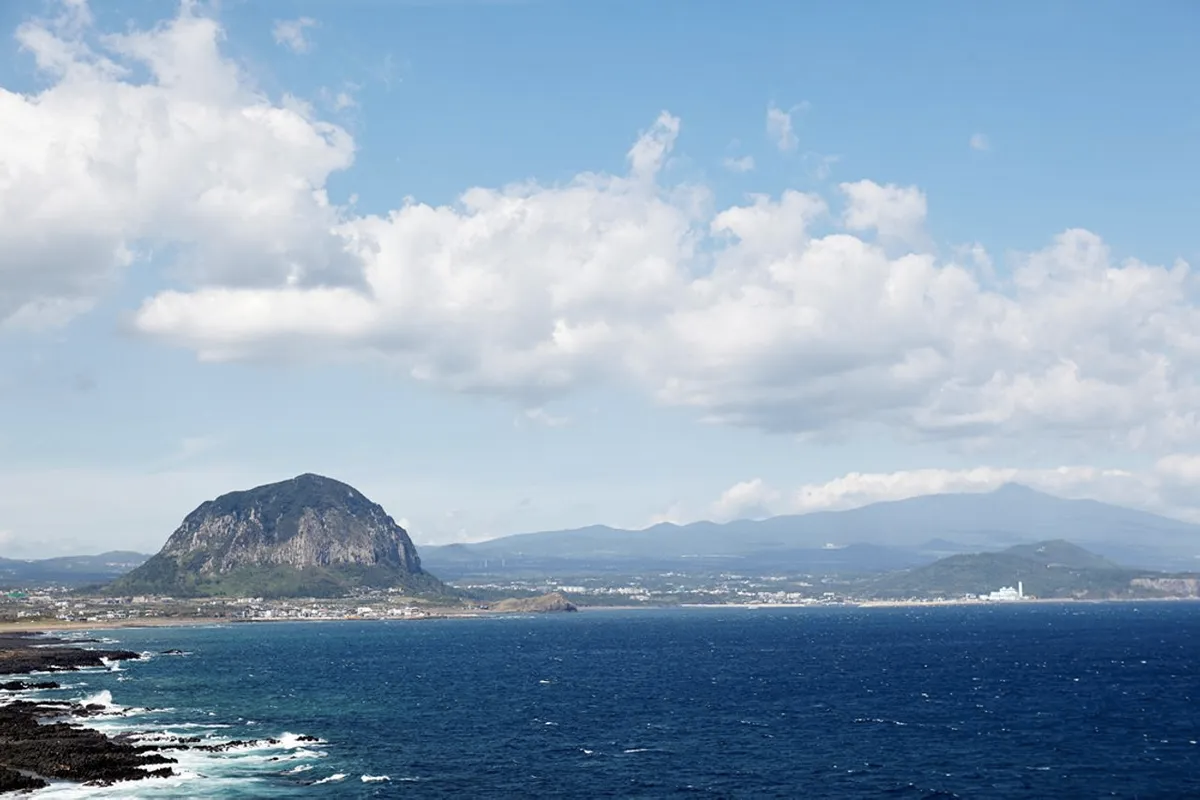 從高空俯瞰濟州島的自然風光，正午陽光下明亮的大海與山景。