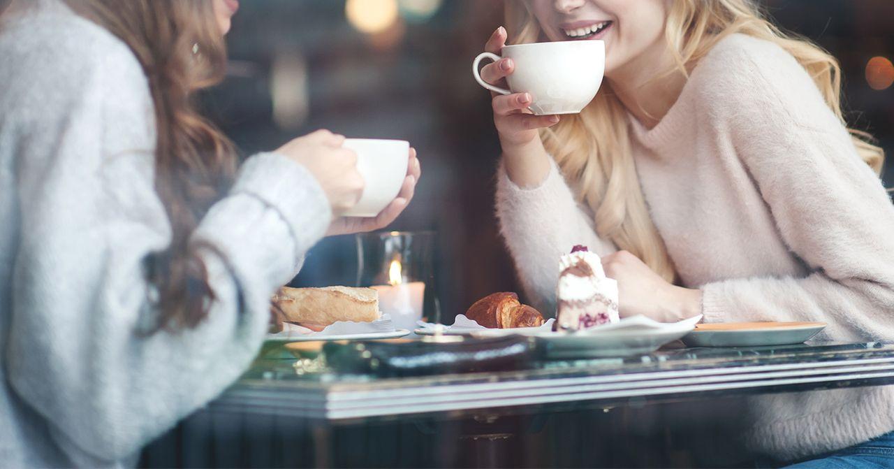 เพื่อนสองคนกำลังสนทนาและดื่มกาแฟในบรรยากาศอบอุ่นในร้านคาเฟ่ ขนมหวานและเครื่องดื่มตั้งอยู่บนโต๊ะไม้ที่มีแสงไฟจากเทียนสร้างบรรยากาศเบาสบาย