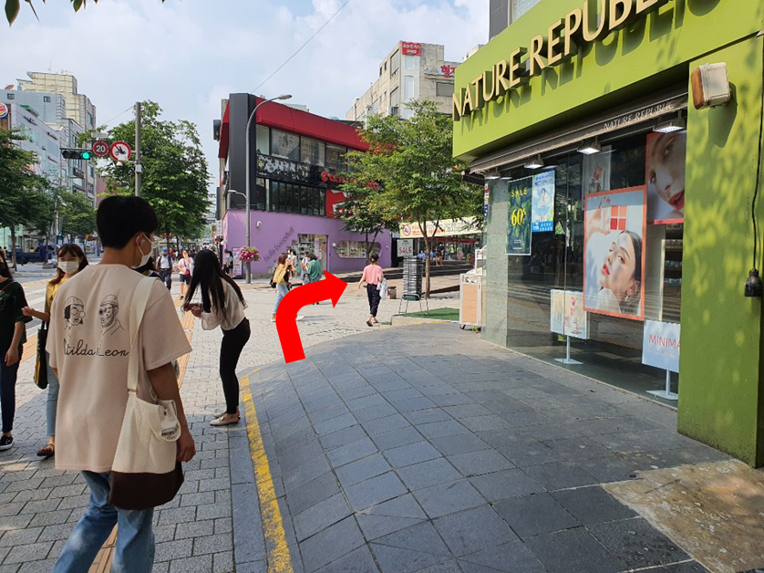 一位顧客在韓國新村街頭行走，享受煦煦陽光，路徑旁邊是一家綠色外觀的Nature Republic店