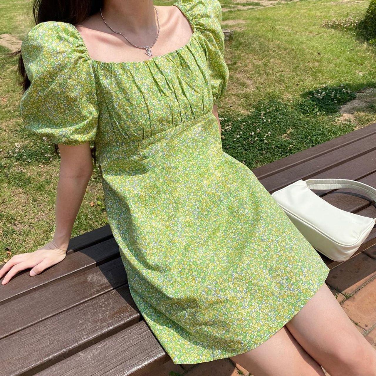 緑色の花柄ミニワンピースを着た女性。公園のベンチに座りながら、白いバッグを横に置いている。