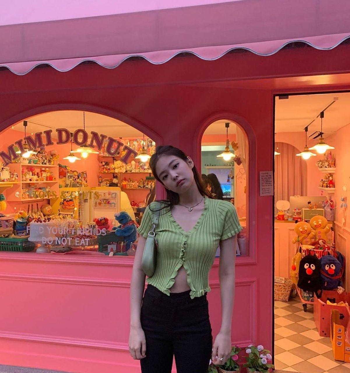 緑のクロップ丈トップスと黒のパンツを着た女性。ピンクの外観の店の前に立っている。