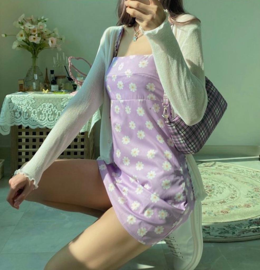 紫の花柄ミニワンピースと白のカーディガンを着た女性。部屋の中でしゃがみ込み、チェック柄バッグを持っている。