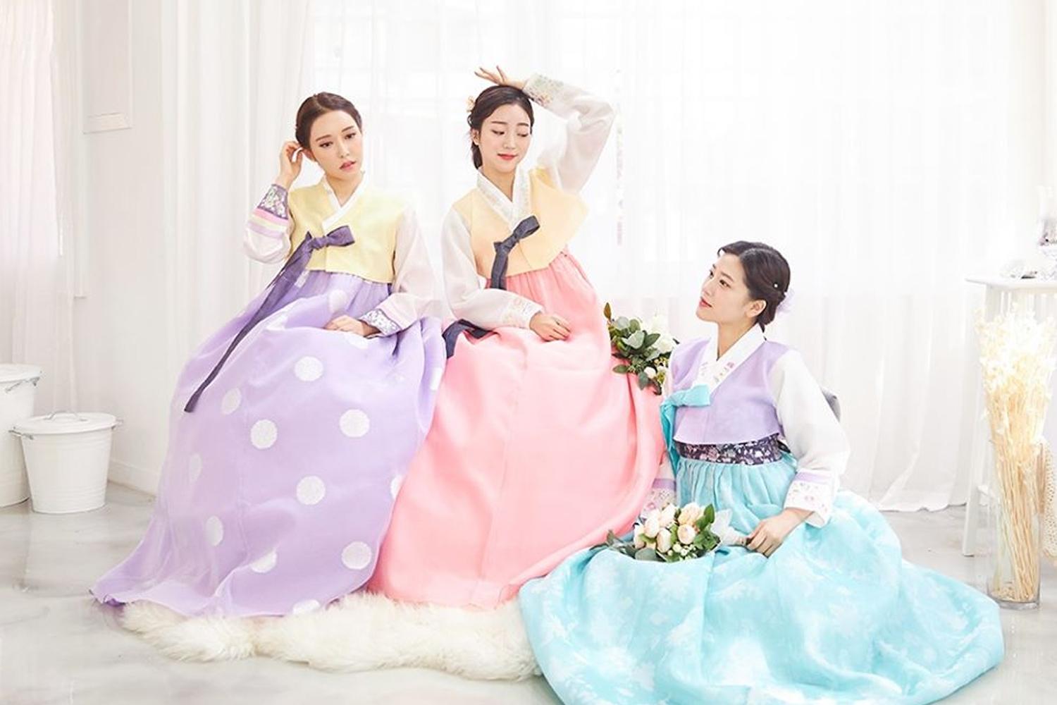 三位穿著色彩鮮豔韓服的女子,坐在室內擺出可愛姿勢拍照。