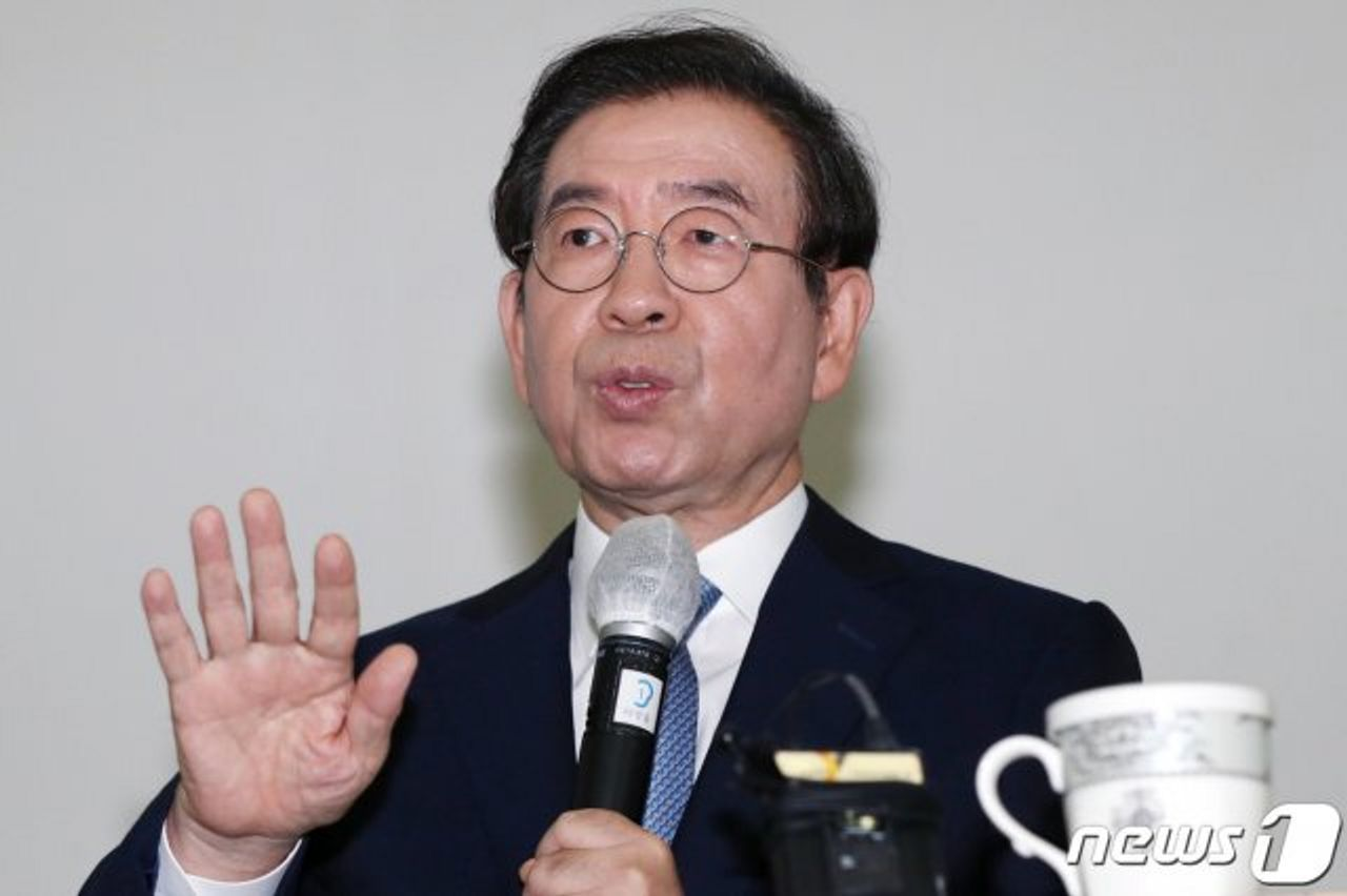 韓國官員在新聞發佈會上揮手背景白色牆壁，話筒和杯子在桌面上