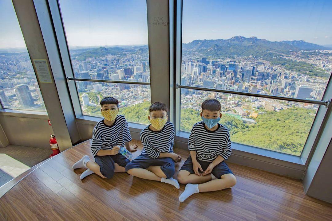 三胞胎在室內觀景區坐著，背景可見首爾城市景觀，享受寧靜的時光。