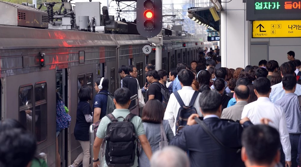 圖片中是一個繁忙的韓國地鐵站臺，乘客們正排隊準備上車或下車，搭乘地鐵的是上班族和平常的乘客，平臺上有紅色的列車信號燈。