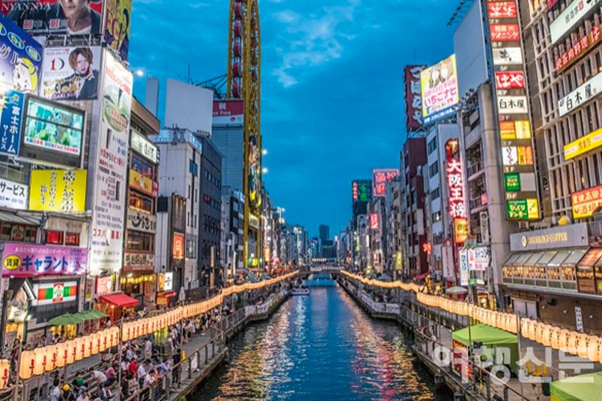 日本大阪的繁華街景，夜晚燈光閃亮，兩側為高聳的寫字樓，途人於水邊行走。圖源：여행신문