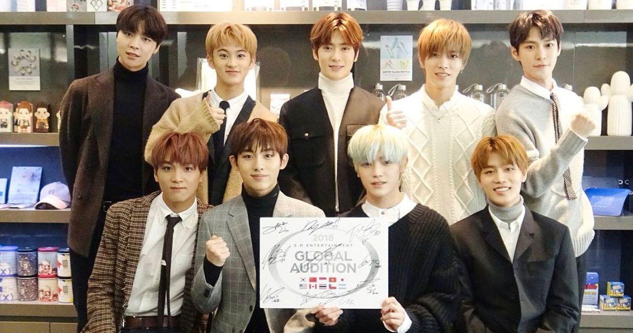 กลุ่มชายหนุ่มในชุดหลากหลายกำลังยืนถ่ายรูปใน SM Entertainment Global Audition ปี 2018 ถือโปสเตอร์ลงนามพร้อมแสดงพลัง