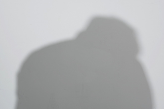 회색 배경에 희미하게 드리워진 사람의 그림자. 고개를 숙인 뒷모습으로 어두운 분위기를 자아냅니다.