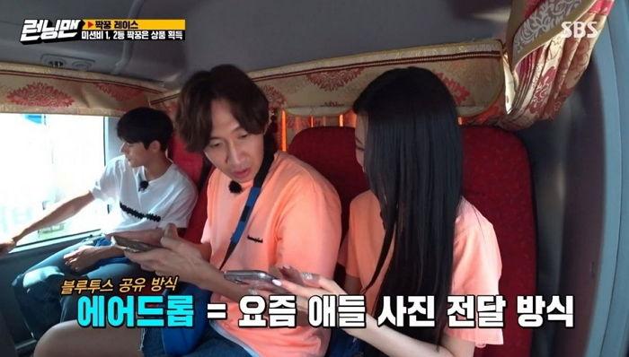 李光洙和張員瑛在《Running Man》的錄影中，正在用手機討論如何用AirDrop分享照片，背景是一輛行駛中的大巴車廂。