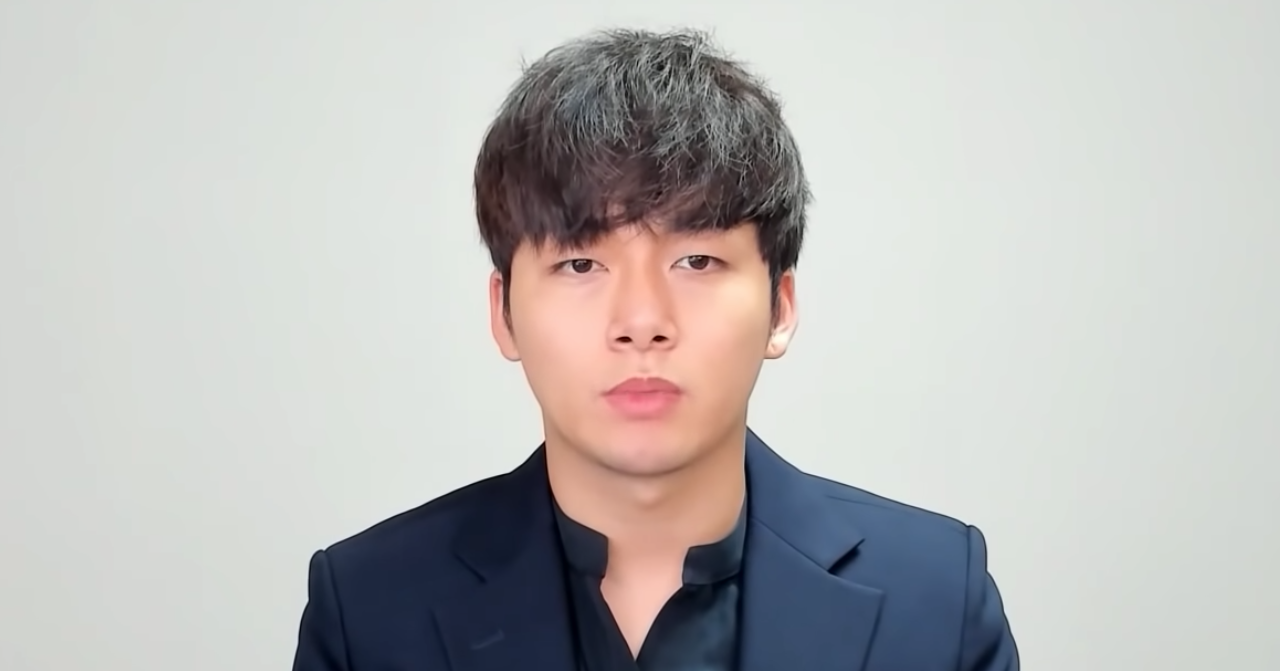 一名南韓男子身穿深色西裝，背景為白色牆面，專注地坐著。