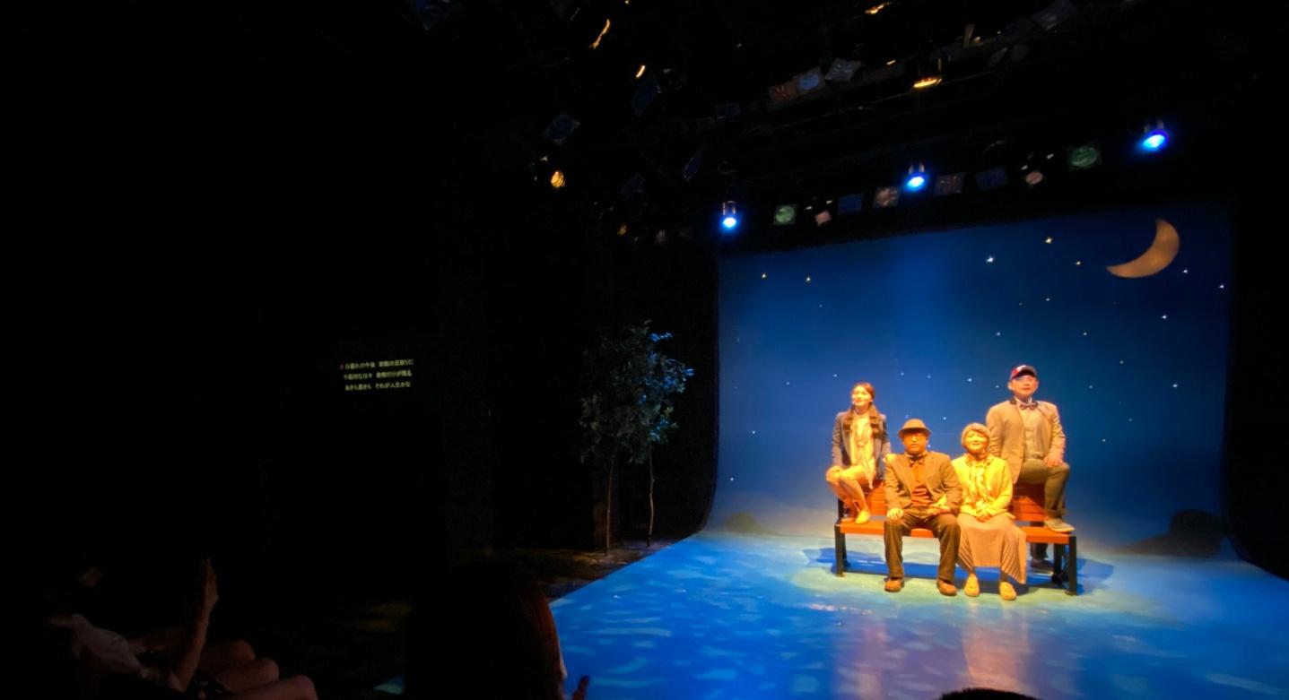 暗い劇場内での舞台公演の様子、夜空の背景に月が浮かぶセット