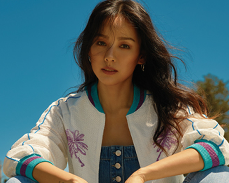 李孝利穿著白色刺繡外套和牛仔裙,坐在户外陽光灑落的地方。