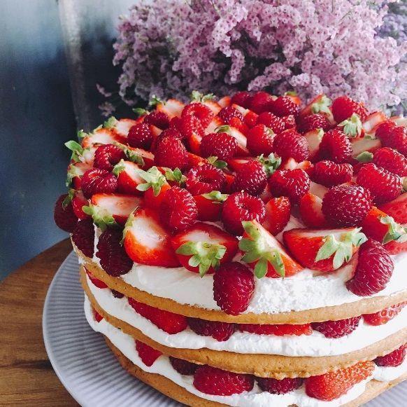 一款滿覆草莓的多層奶油蛋糕，以優雅的姿態展示在桌上，背景的紫色花朵增添點綴。