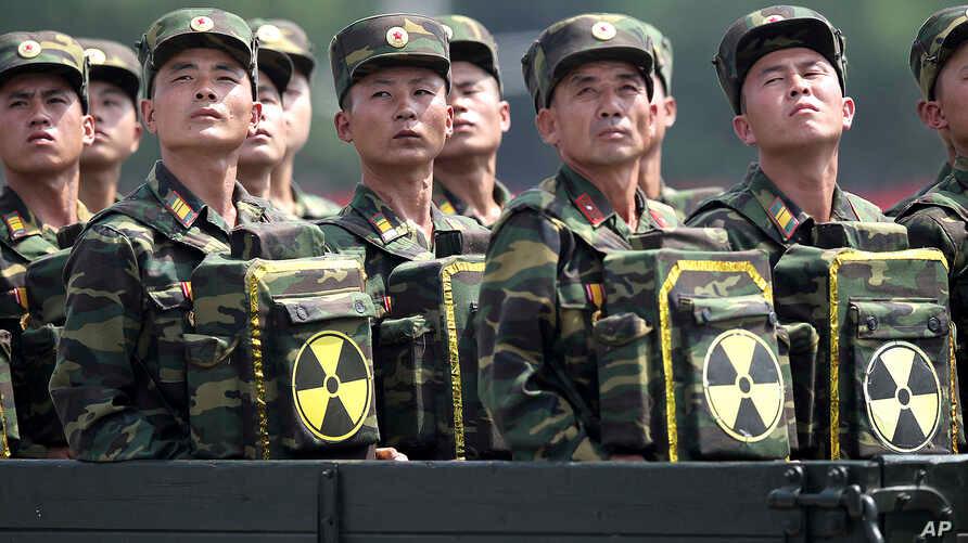 ทหารเกาหลีเหนือในชุดลายพรางยืนต่อแถวในขบวนพาเหรด แต่ละคนถือกล่องยุทธวิธีพร้อมสัญลักษณ์นิวเคลียร์ แสดถึงการรวมตัวของกองทัพและพลังทางการทหาร