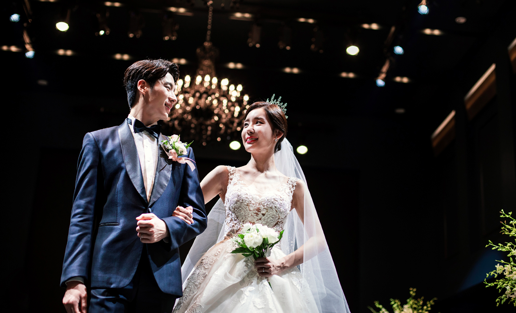 在豪華婚禮場地中的新郎和新娘，新娘穿著禮服，手持捧花，周圍裝飾著水晶吊燈和花束。