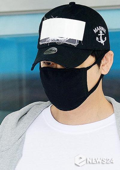 フードをかぶり、黒いマスクをした男性が空港に到着する様子。黒いキャップには海軍のアンカーの刺繍があり、白いTシャツの上にグレーのパーカーを着ている。ニュース24のロゴが右下に配置されている。