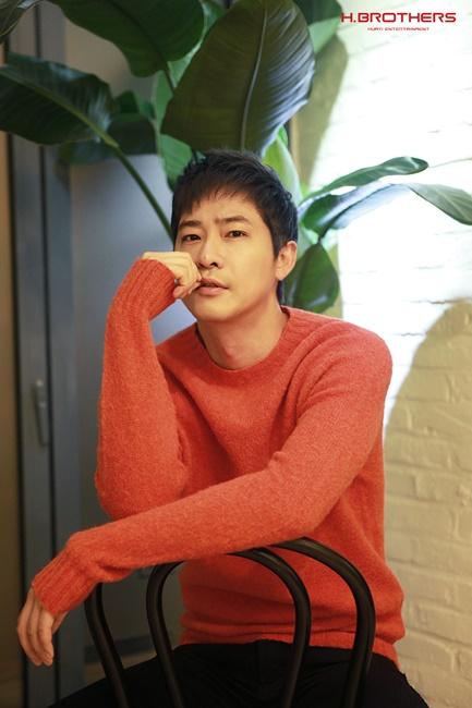 オレンジ色のセーターを着た男性が黒い椅子に腰掛けている。背後には大きな観葉植物が置かれており、H.Brothersのロゴが右上に表示されている。室内のレンガ調の壁と白っぽい光がアクセントとなっている。