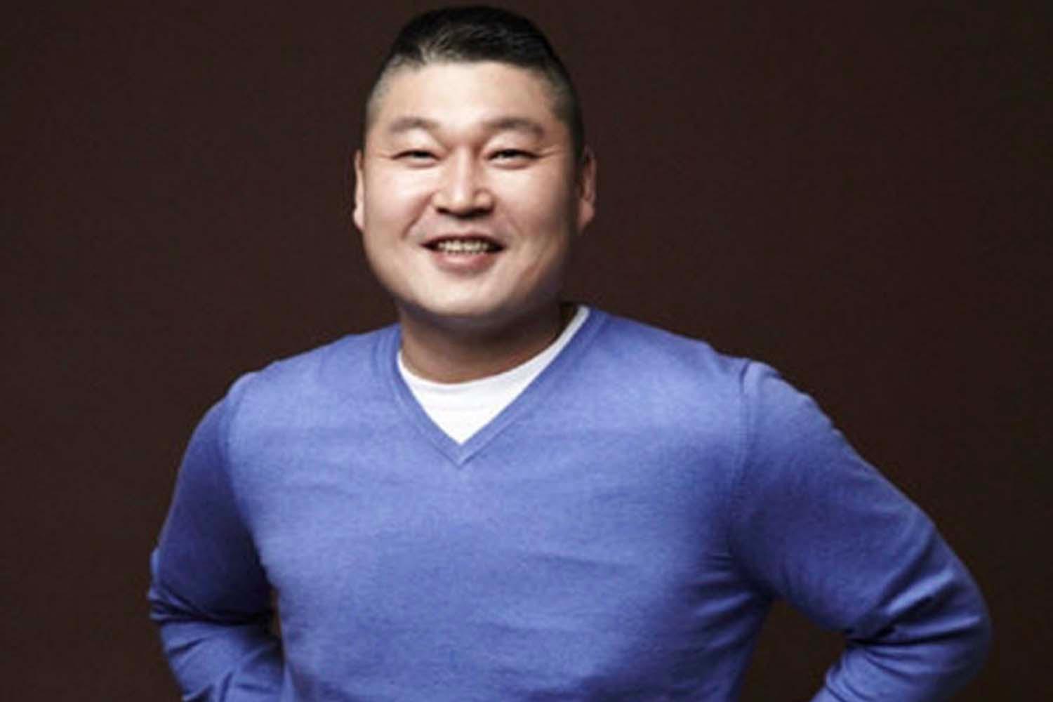 คังโฮดงสวมเสื้อสเวตเตอร์สีฟ้าเข้มยืนโพสท่าพร้อมพื้นหลังสีน้ำตาลเข้ม